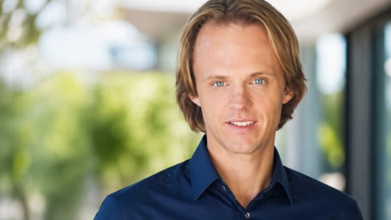 Editorial portrait representing David Wilcock died topic, showing a UFO researcher in a dim study with books, notes, and cosmic imagery tied to his legacy