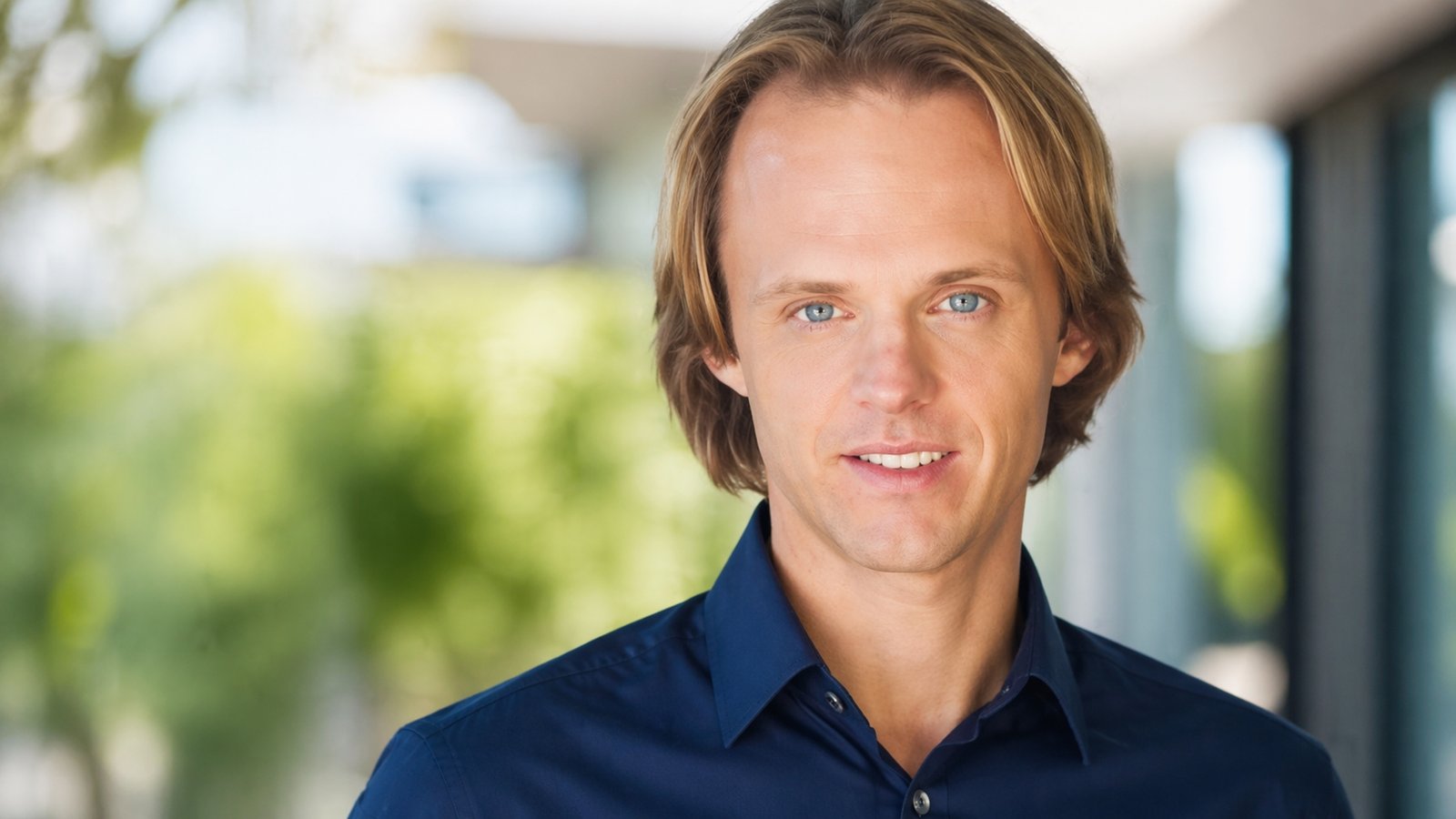 Editorial portrait representing David Wilcock died topic, showing a UFO researcher in a dim study with books, notes, and cosmic imagery tied to his legacy
